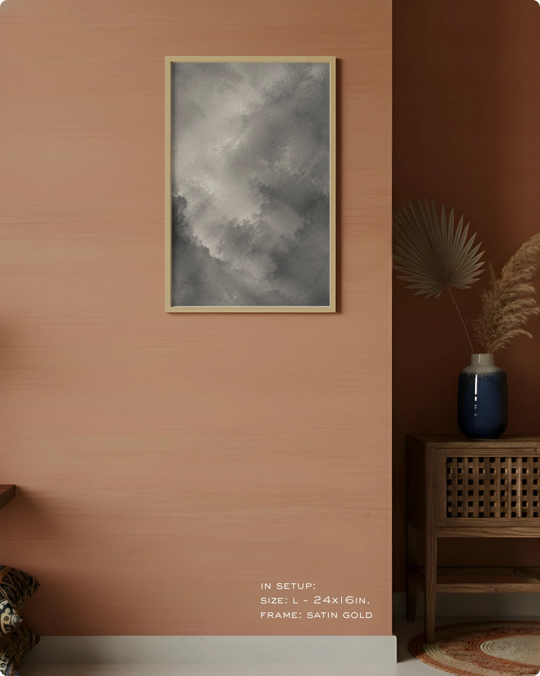 A framed wall art print with a black and white stormy sky design, hung on a peach-colored wall in a styled interior setting with a wooden side table, blue vase, and dried palm leaves.