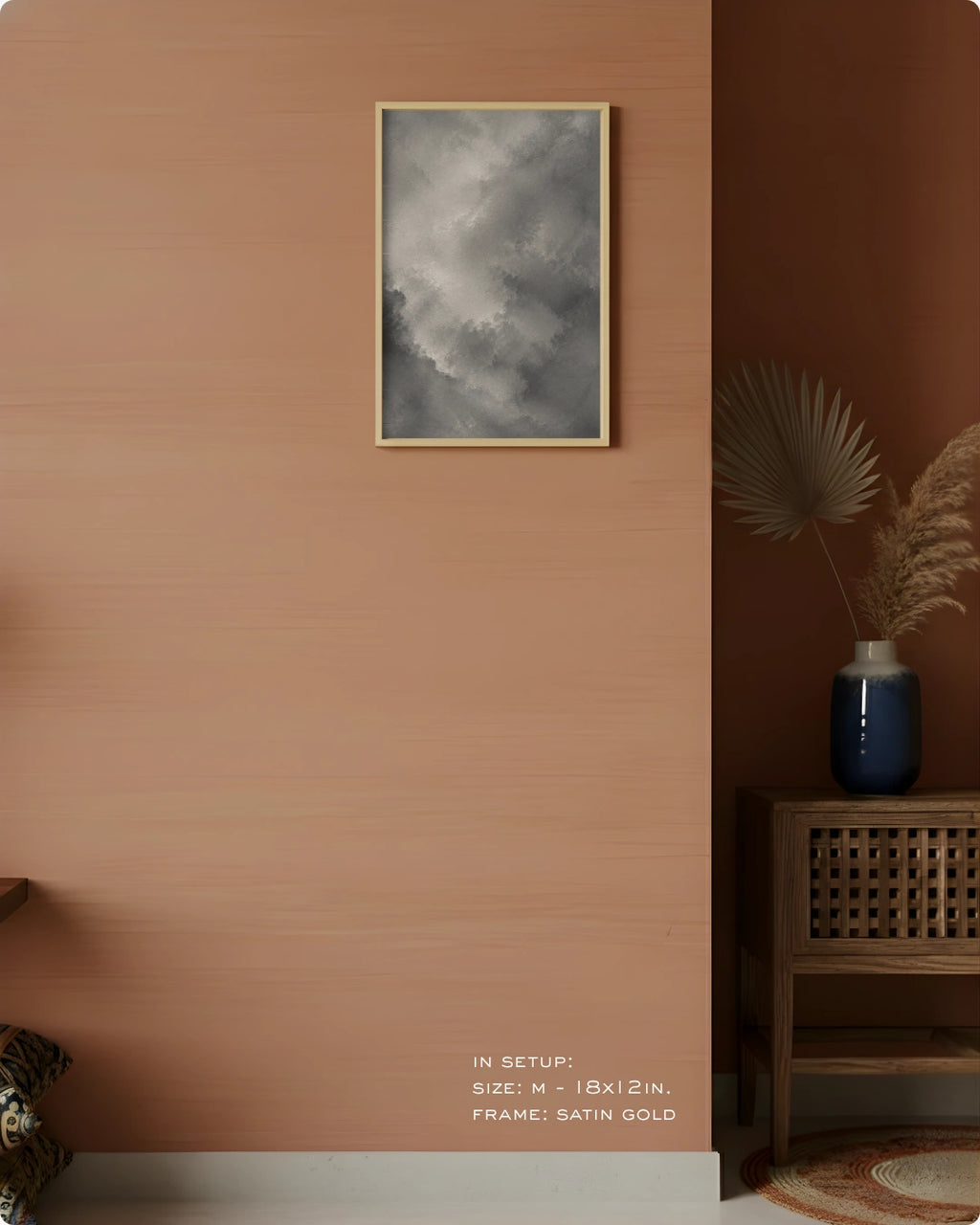 A framed wall art print showing a cloudy gray sky, hung on a warm terracotta-colored wall in a styled interior setting with a wooden side table, blue vase, dried palm leaves, and small text indicating size and frame details.