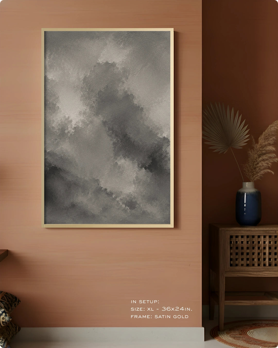 A framed wall art print with a gray and white abstract cloudy design, displayed on a warm peach-colored wall in a styled interior setting with a wooden side table and vase.