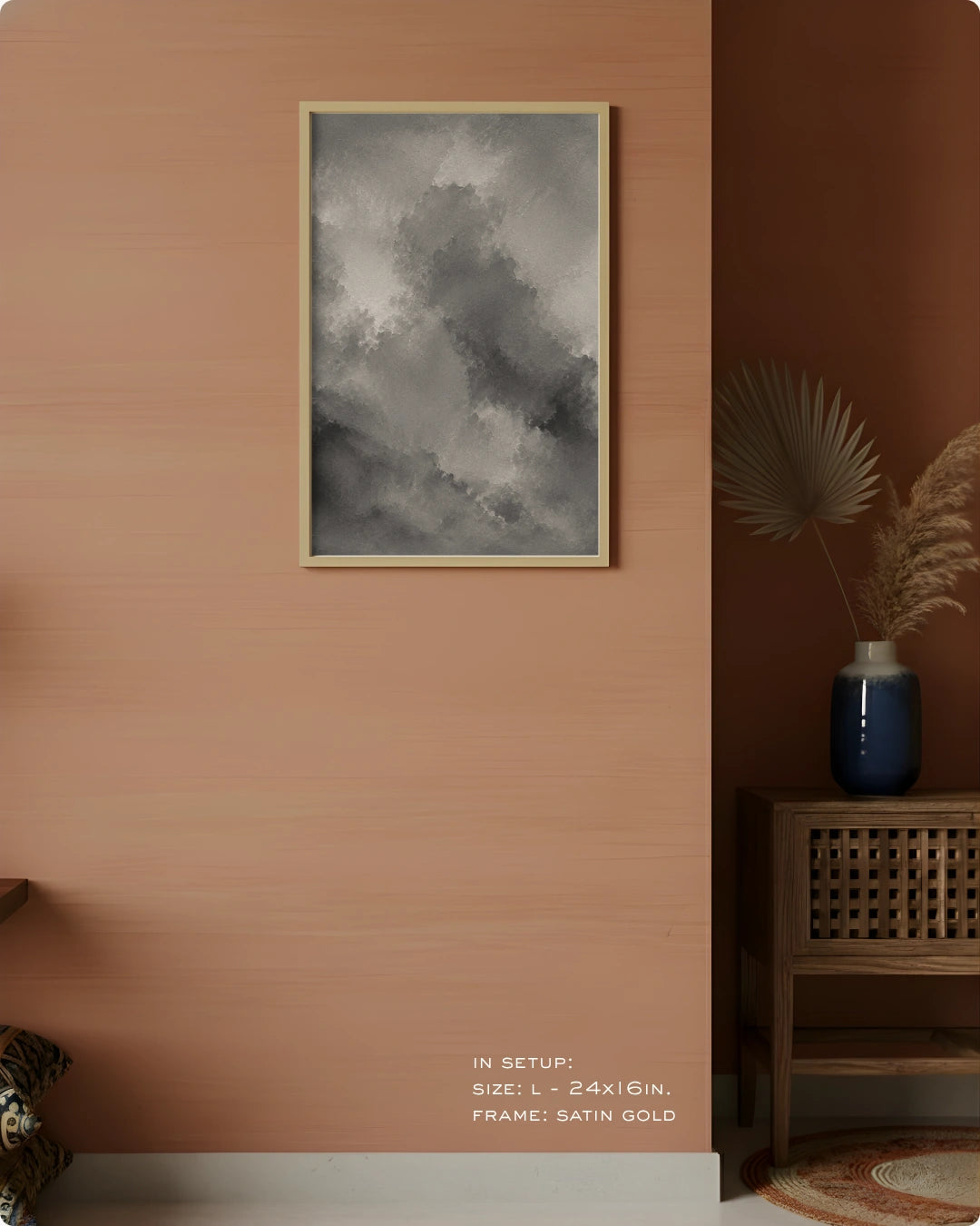 A framed wall art print with a gray and white cloudy abstract design hangs on a warm peach-pink wall in a styled interior. A wooden side table with a blue vase and dried palm leaves sits to the right, and small text at the bottom reads 'IN SETUP: SIZE L = 24x6IN.
