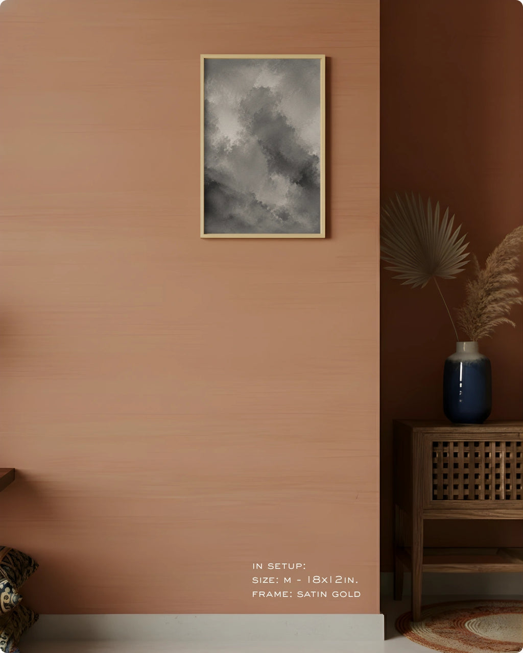 A framed wall art print featuring a dramatic gray and black cloudy sky, hung on a warm terracotta-colored wall in a styled interior setting with a wooden side table, blue vase, and dried palm leaves.