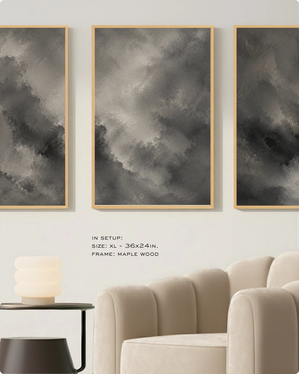 Three vertical framed wall art panels with abstract gray and black cloud-like brushstrokes hang on a light wall above a beige armchair and side table. Small text reads: IN SETUP: SIZE: XL - 36x24in, FRAME: MAPLE WOOD.