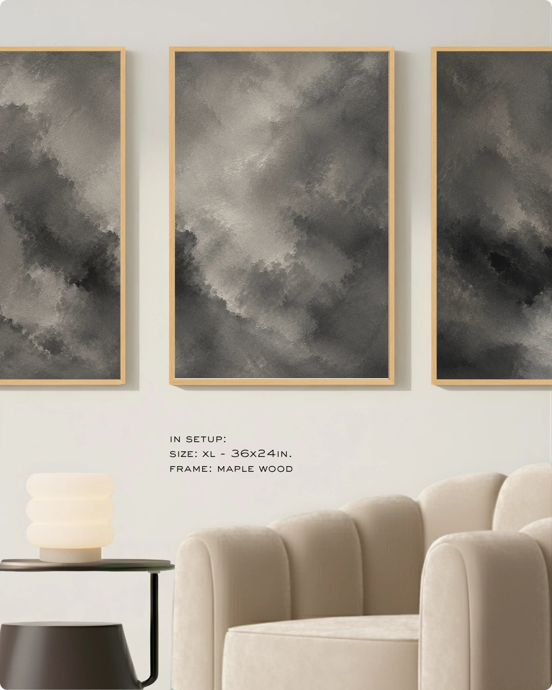 Three vertical framed wall art panels with abstract gray and black cloud-like brushstrokes hang on a light wall above a beige armchair and side table. Small text reads: IN SETUP: SIZE: XL - 36x24in, FRAME: MAPLE WOOD.