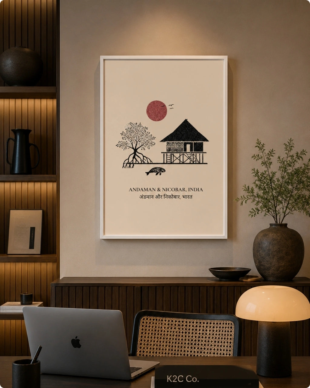 A framed minimalist art print hangs on a beige wall in a modern, warmly lit home office. The print features a black line drawing of a house, tree, and bird under a red sun, with the text 'ANDAMAN & NICOBAR, INDIA' and 'aspen tree forest'.