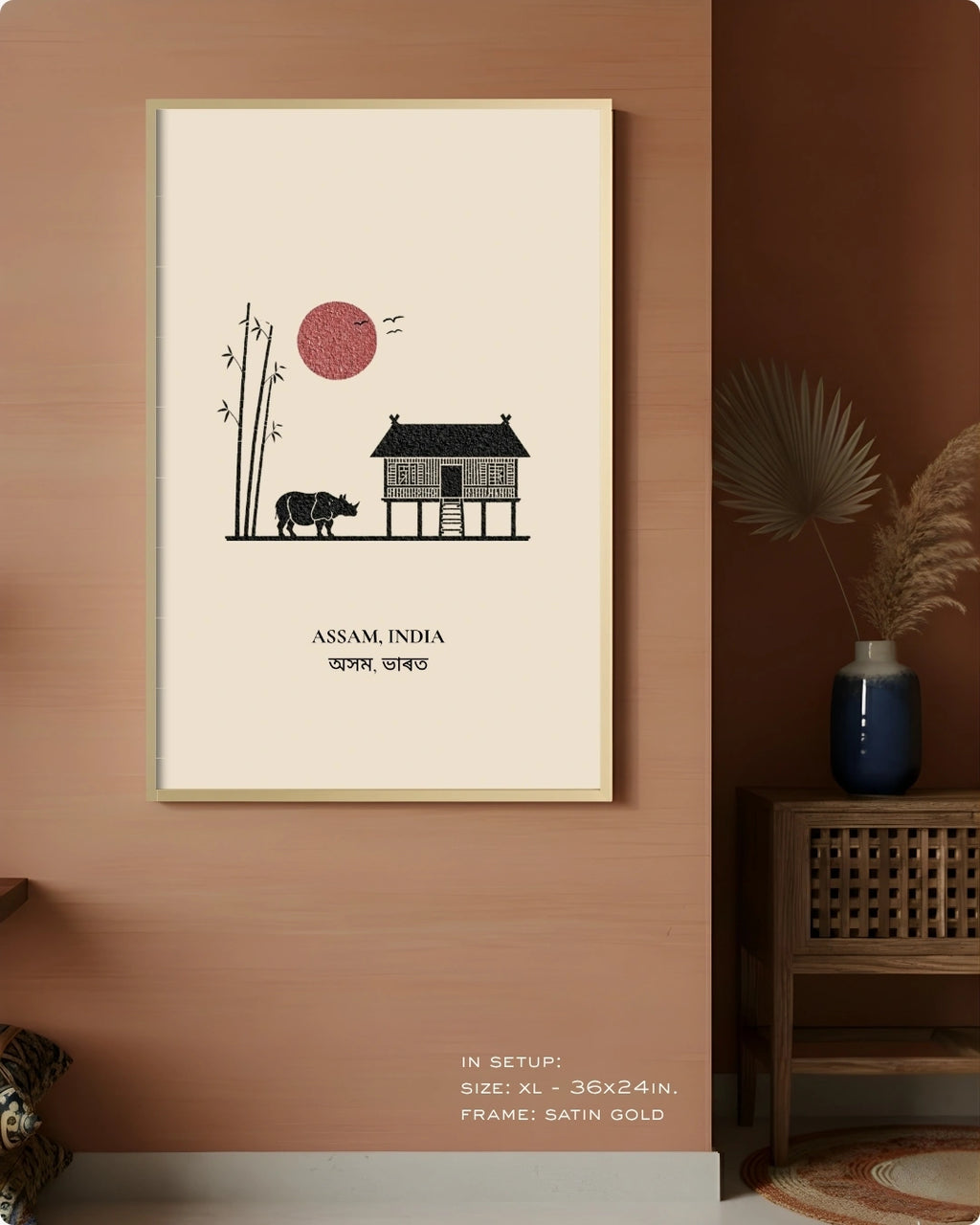 A framed wall art print hangs on a terracotta-colored wall in a styled interior. The print features a minimalist illustration of a house, a cow, bamboo, and a red sun with the text 'ASSAM, INDIA' and Devanagari text below.