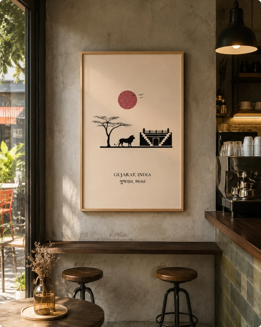 A framed art print featuring a minimalist black silhouette of a Gujarat landscape with a red sun on a cream background, hanging on a textured gray wall in a cafe setting.