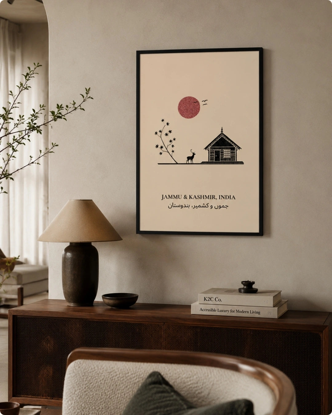 A framed minimalist art print featuring a small house, a red sun, and text reading 'JAMMU & KASHMIR, INDIA' with Arabic text below, displayed on a beige wall above a dark wood console table with a lamp and books.
