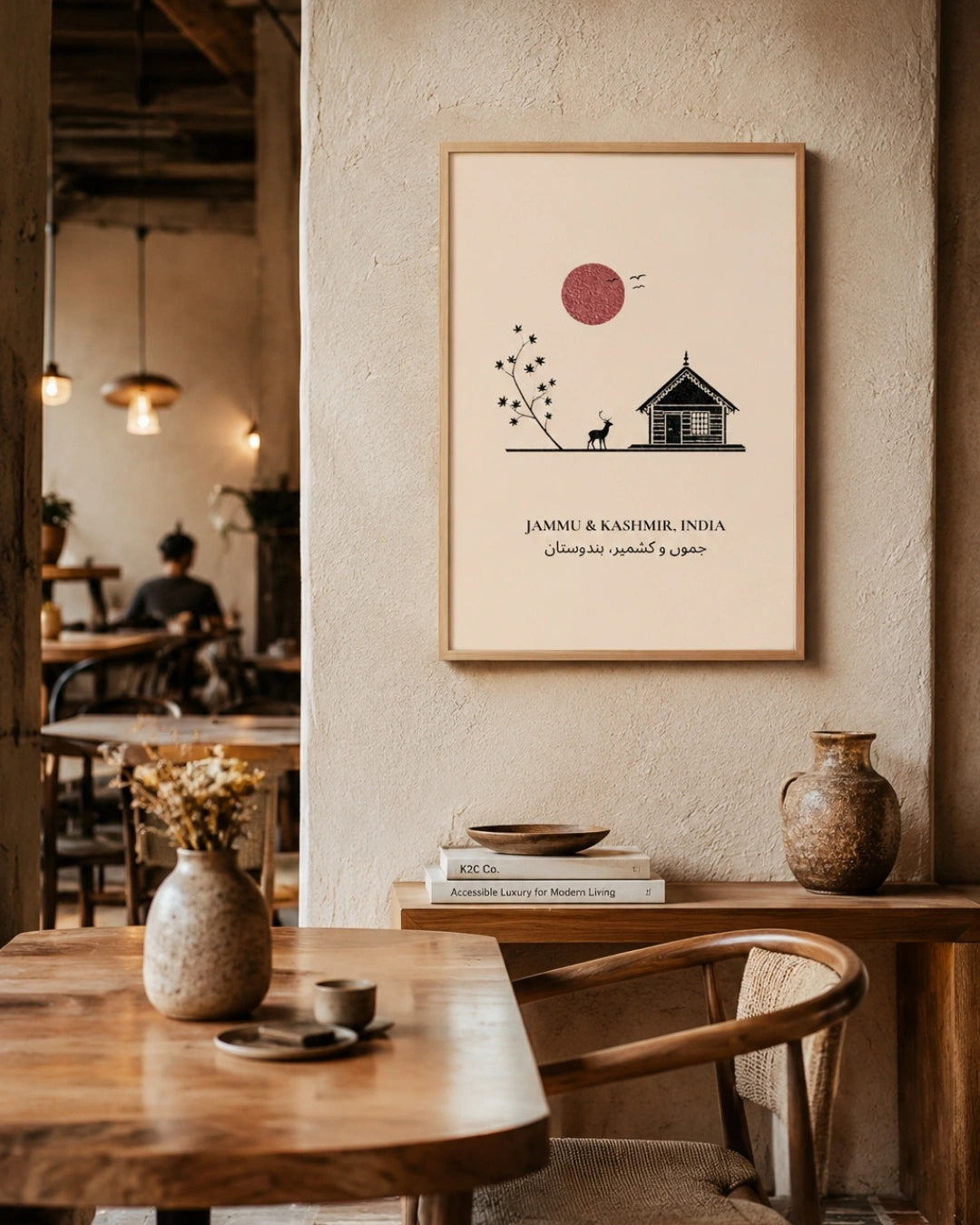 A framed minimalist art print featuring a small house, a tree, a red sun, and the text 'JAMMU & KASHMIR, INDIA' in English and Urdu, displayed on a textured cream wall in a warm, rustic cafe interior.