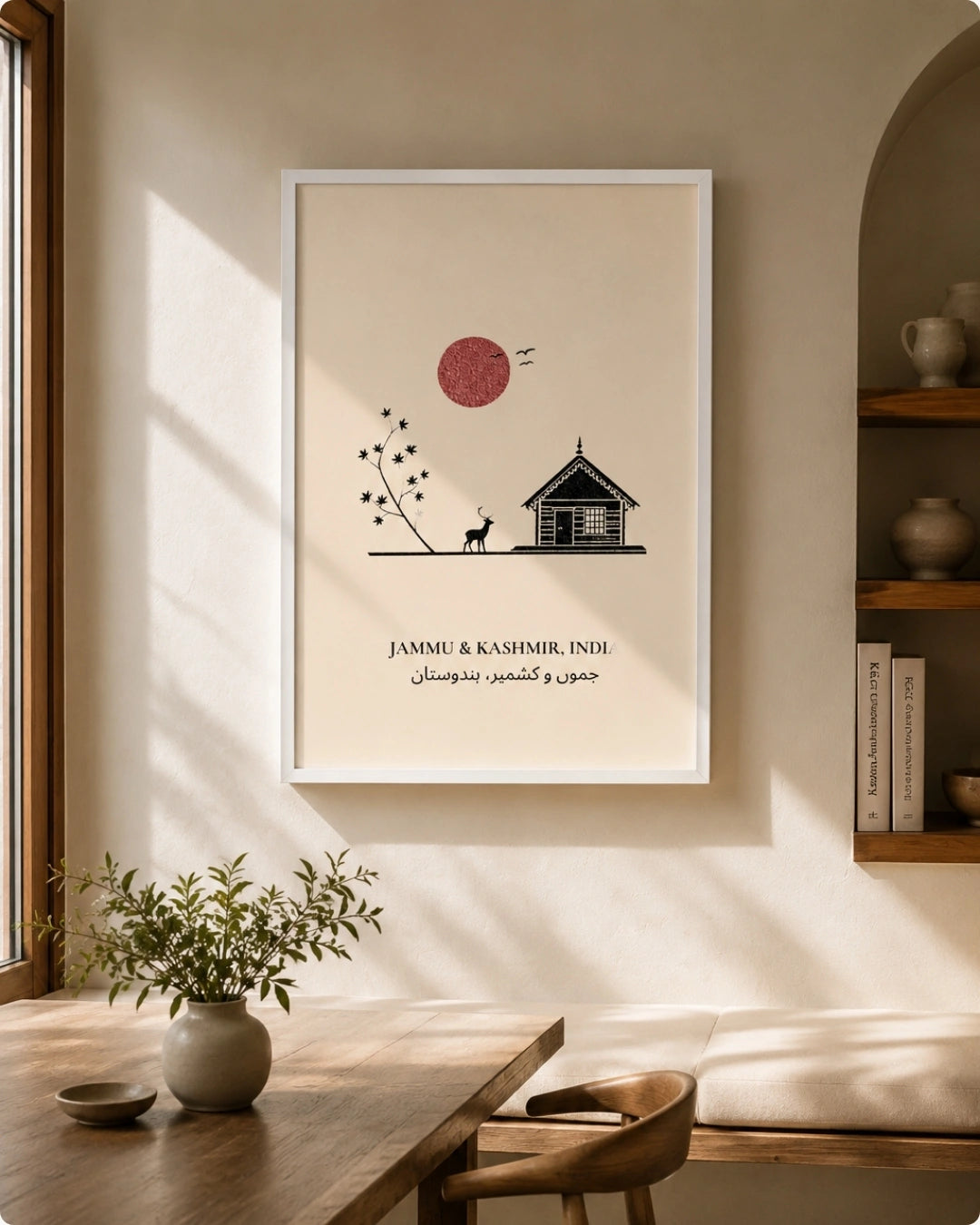 A framed minimalist art print hangs on a warm beige wall in a sunlit room. The print shows a small house, a tree, a dog, and a red sun with the text 'JAMMU & KASHMIR, INDI' and Arabic script below.