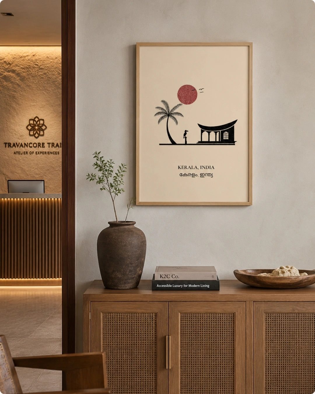 A framed travel art print featuring Kerala's Punalur Palace and a palm tree under a red sun, styled on a light gray wall above a wooden cabinet in a modern interior setting.