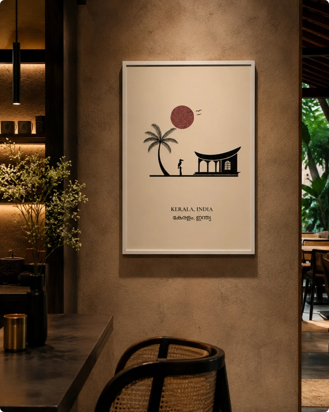 A framed minimalist art print featuring a black silhouette of a house and palm tree under a red sun on a cream background, with the text 'KERALA, INDIA' and Malayalam script, displayed on a textured beige wall in a modern interior setting.