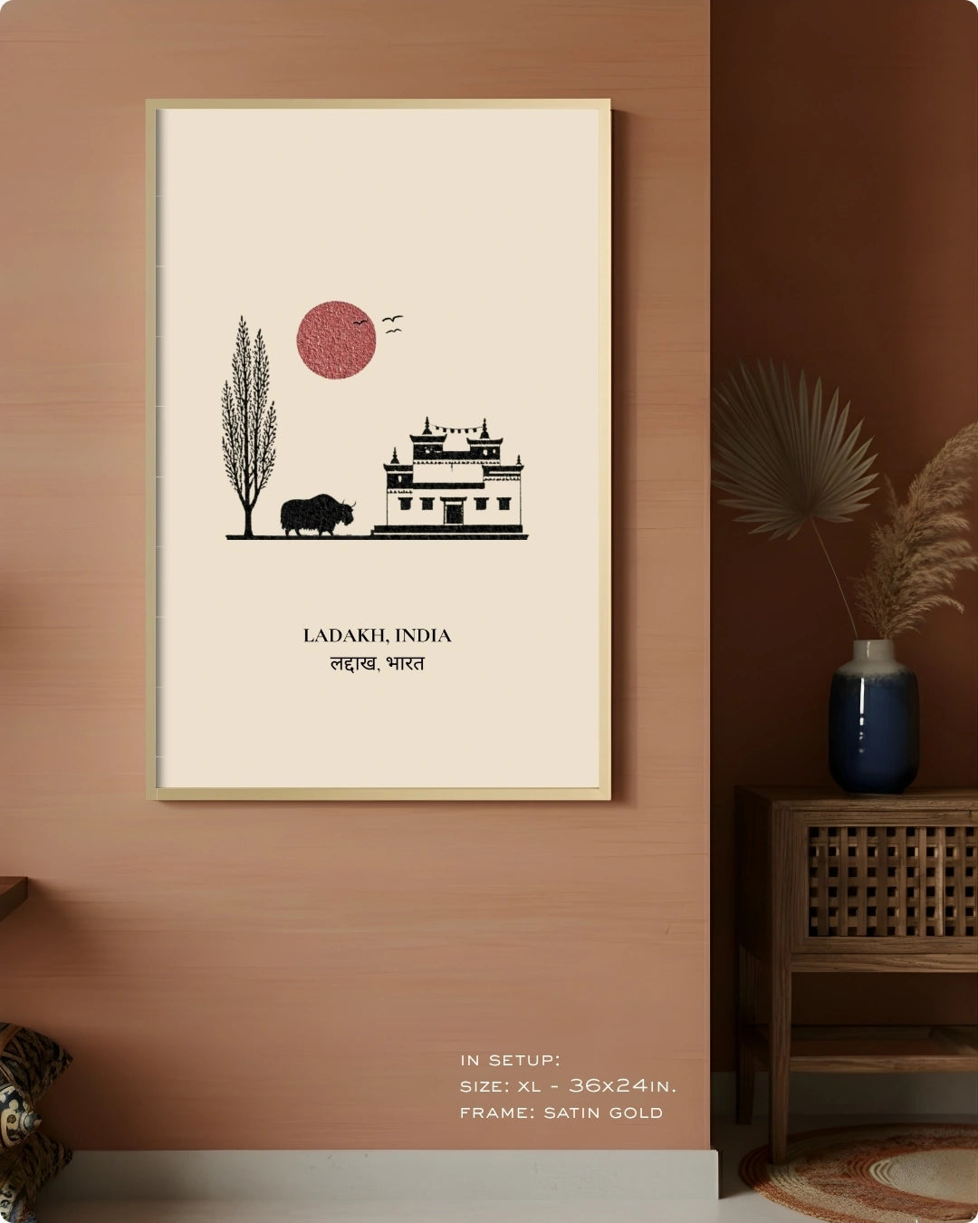 A framed wall art print featuring a minimalist illustration of Ladakhi architecture, a tree, a buffalo, and a red sun on an off-white background, hung on a terracotta-colored wall in a styled interior setting.