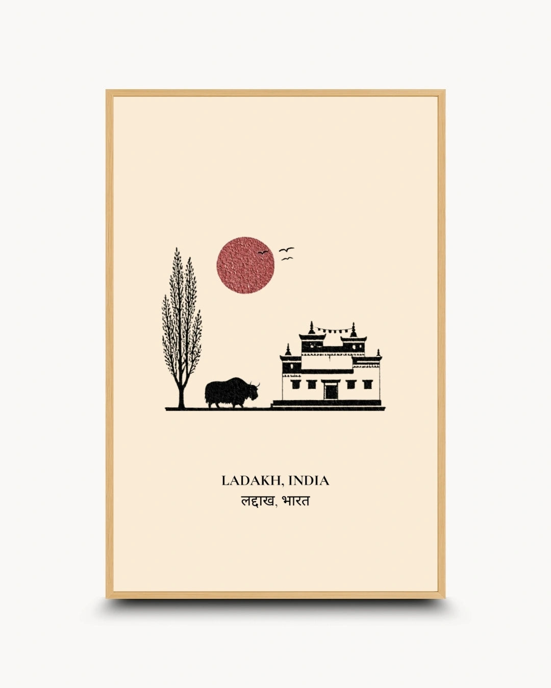 A framed minimalist art print of Ladakh, India, featuring a black silhouette of a traditional building, a tree, a buffalo, and a red sun on a cream background, enclosed in a light wood frame.