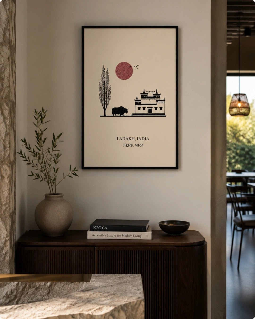 A framed minimalist art print featuring a red sun, a tree, a buffalo, and a house with the text 'LADAKH' displayed on a wall above a dark wooden console table with a vase and books.