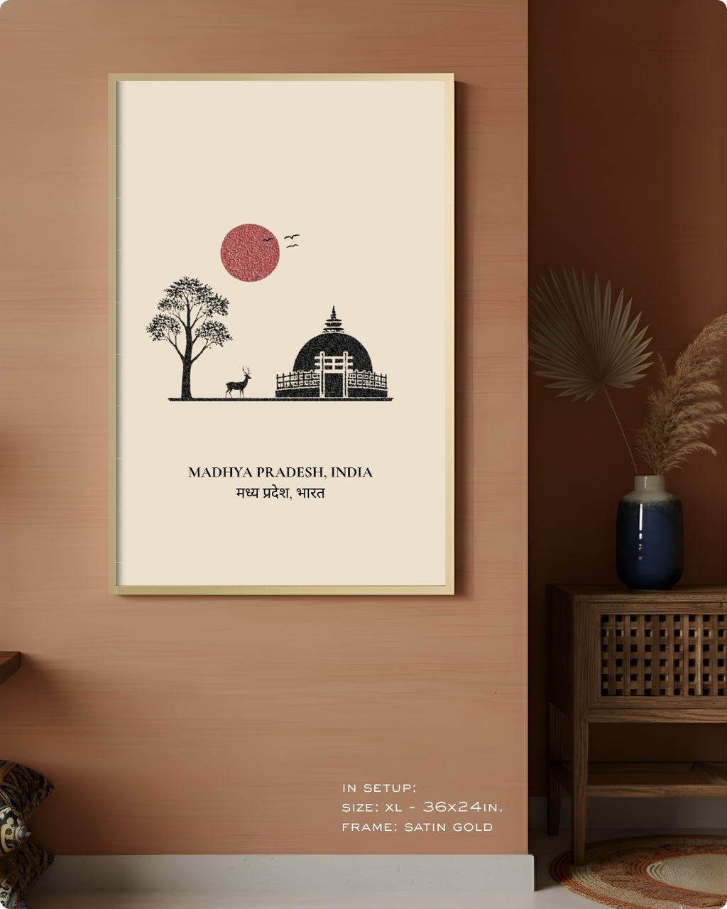 A framed wall art print hangs on a terracotta-colored wall in a styled interior. The print features a minimalist beige background with a black line drawing of a domed building, a tree, a deer, and a pink sun, with the text 'MADHYA PRADESH, INDIA' below.