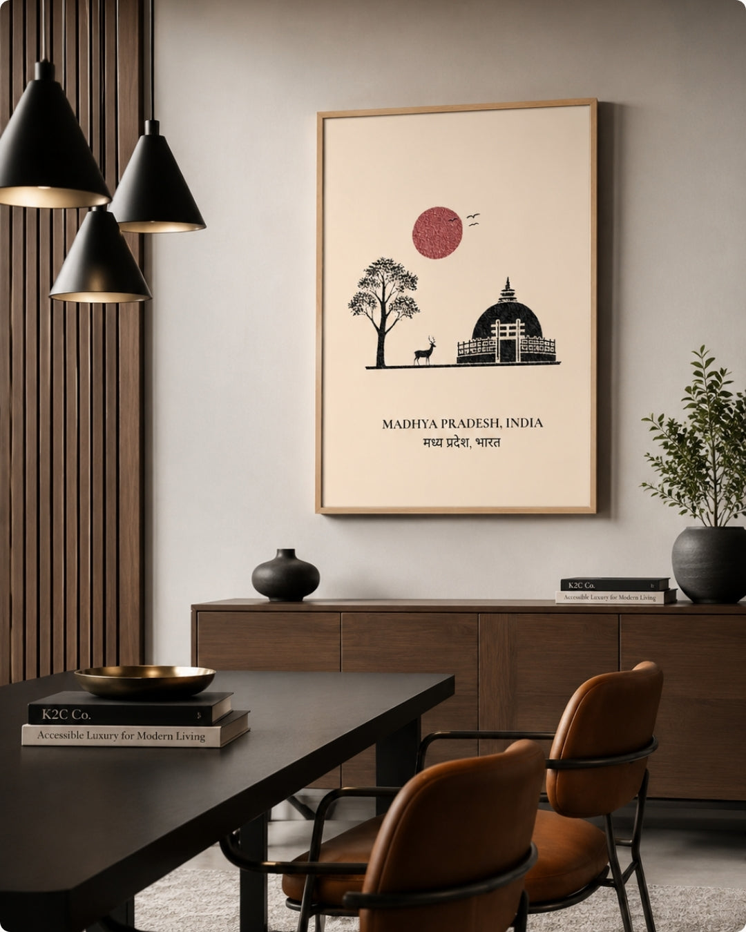 A framed wall art print featuring a minimalist black line drawing of a tree, a domed building, and a red sun on a cream background, with the text 'MADHYA PRADESH, INDIA' below. The print is displayed in a modern dining room setting above a wooden credenza.
