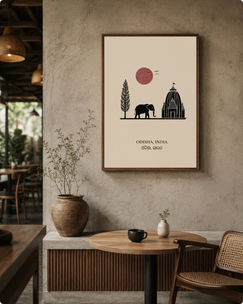 A framed art print featuring a minimalist black line drawing of an elephant, a tree, and a domed temple under a pink sun on a cream background, hanging on a textured gray wall in a cafe setting.