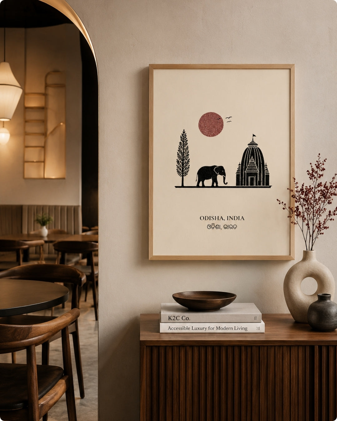 A framed minimalist art print featuring a black silhouette of an elephant, a tree, and a temple with a red sun, displayed on a beige wall in a modern, warm-toned interior setting.