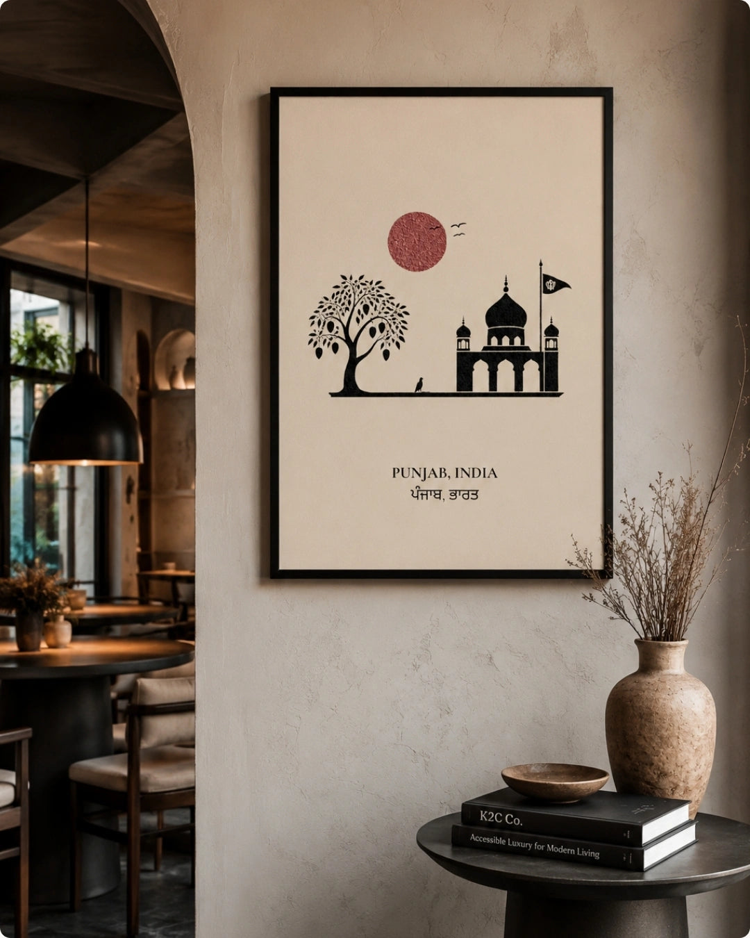 A framed minimalist art print featuring a black silhouette of a tree and a building under a dusty pink sun, with the text 'PUNJAR, INDIA' and 'April 2022', displayed on a textured beige wall in a modern interior setting.