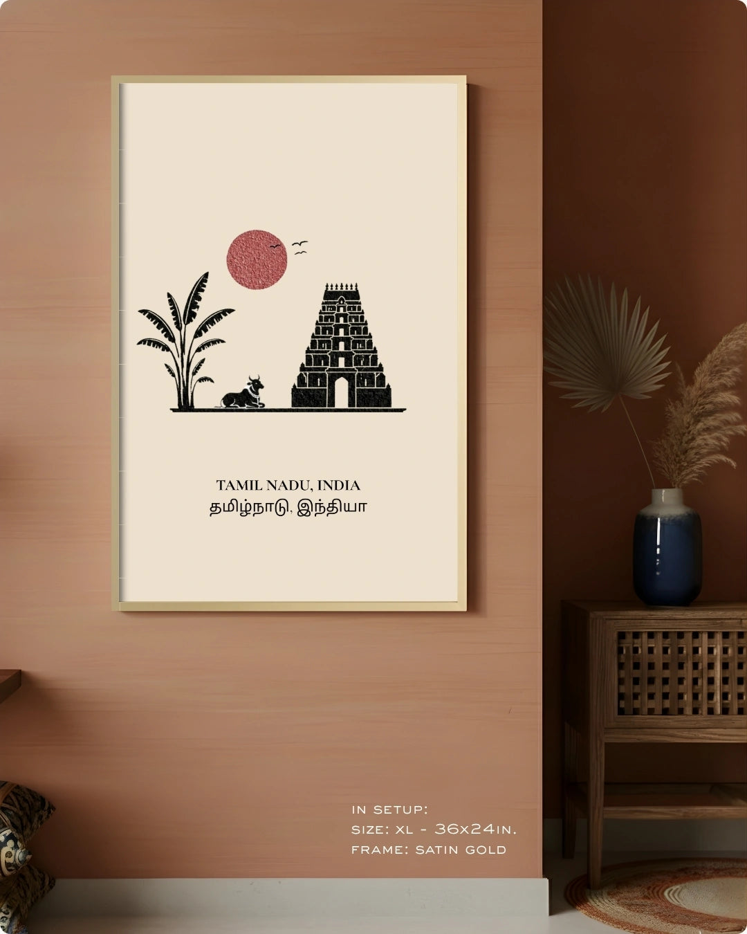 A framed vertical wall art print hangs on a terracotta-colored wall in a styled interior. The print features a minimalist black illustration of a Tamil Nadu temple tower with a pink sun and plant silhouette on a warm cream background, surrounded by a thin gold frame.