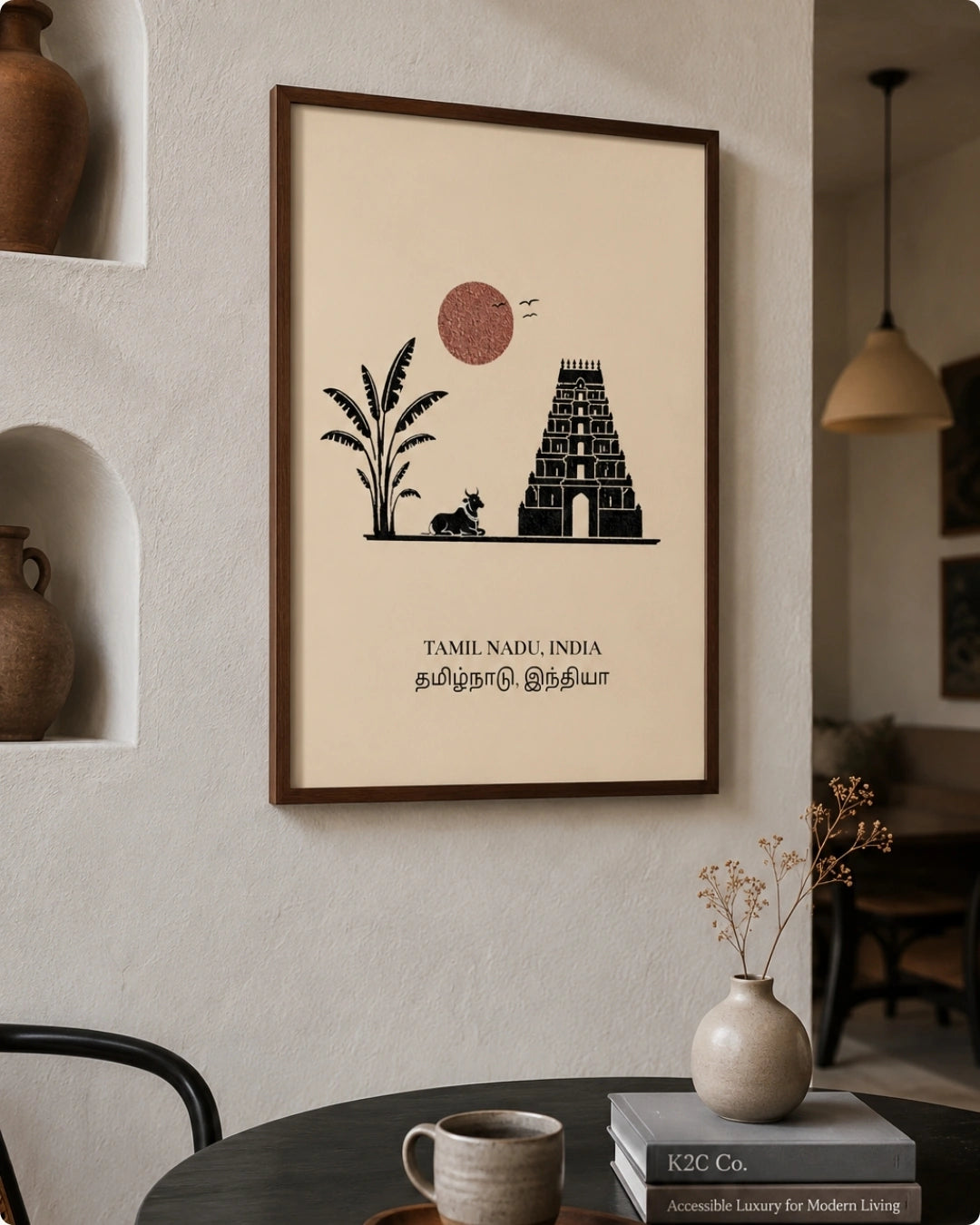 A framed minimalist Tamil Nadu art print hangs on a textured white wall in a styled interior setting. The print features a black silhouette of a temple tower, a palm tree, a small monkey, and a pinkish sun on a cream background, enclosed in a dark brown frame.