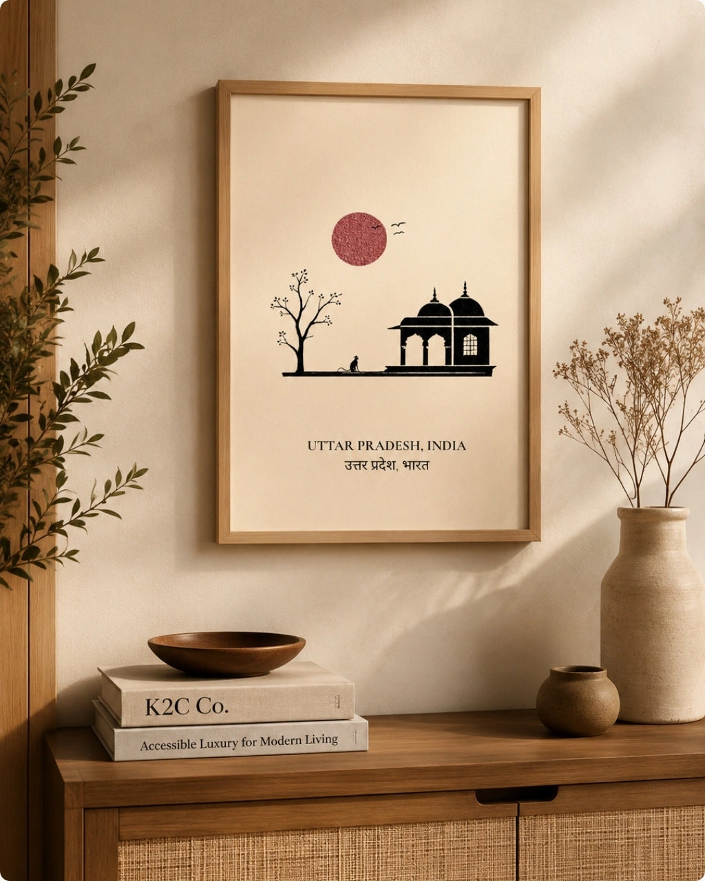 A framed minimalist art print featuring a black silhouette of a domed building and tree under a dusty pink sun on a cream background, hanging on a beige wall above a wooden console table with books and dried flowers.