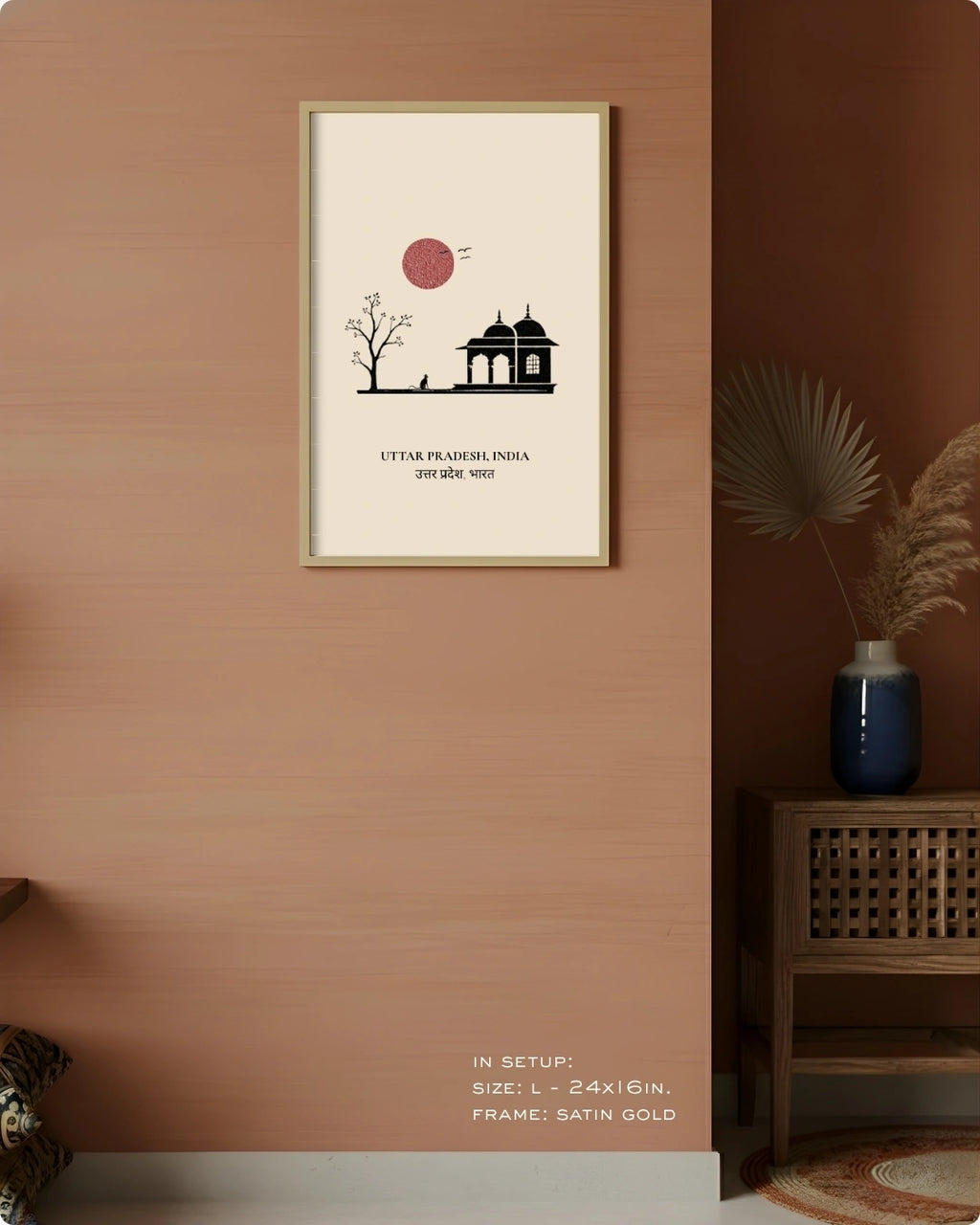A framed minimalist art print featuring a black silhouette of a building and tree under a red sun on a cream background, hung on a warm terracotta wall in a styled interior setting.