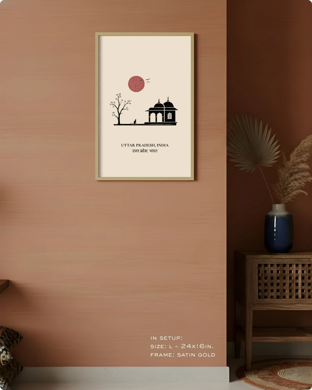 A framed minimalist art print featuring a black silhouette of a building and tree under a red sun on a cream background, hung on a warm terracotta wall in a styled interior setting.