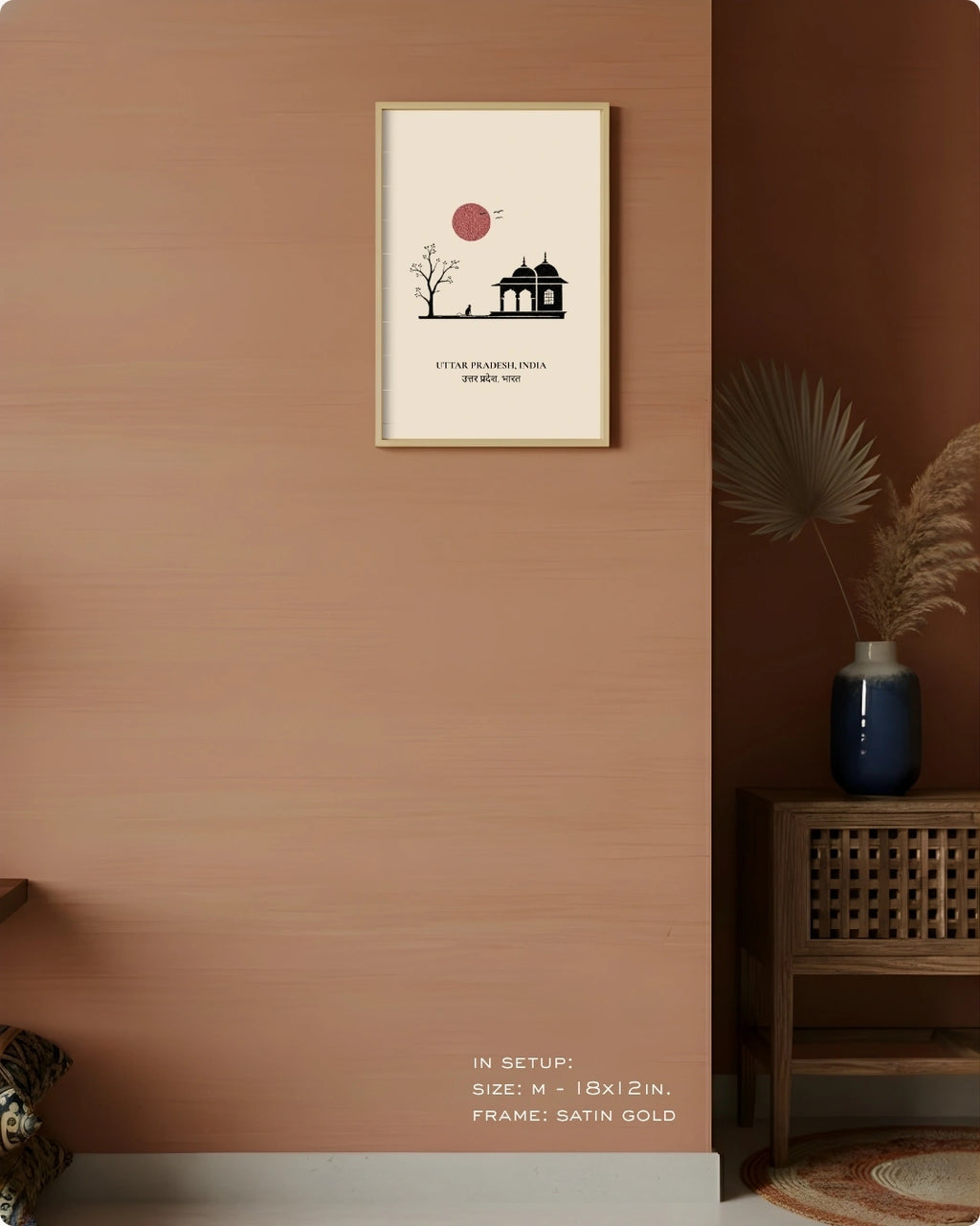 A framed minimalist art print hangs on a warm terracotta-colored wall in a styled interior setting. The print features a cream background with a black line drawing of a pavilion and tree under a muted red sun, surrounded by a thin satin gold frame.