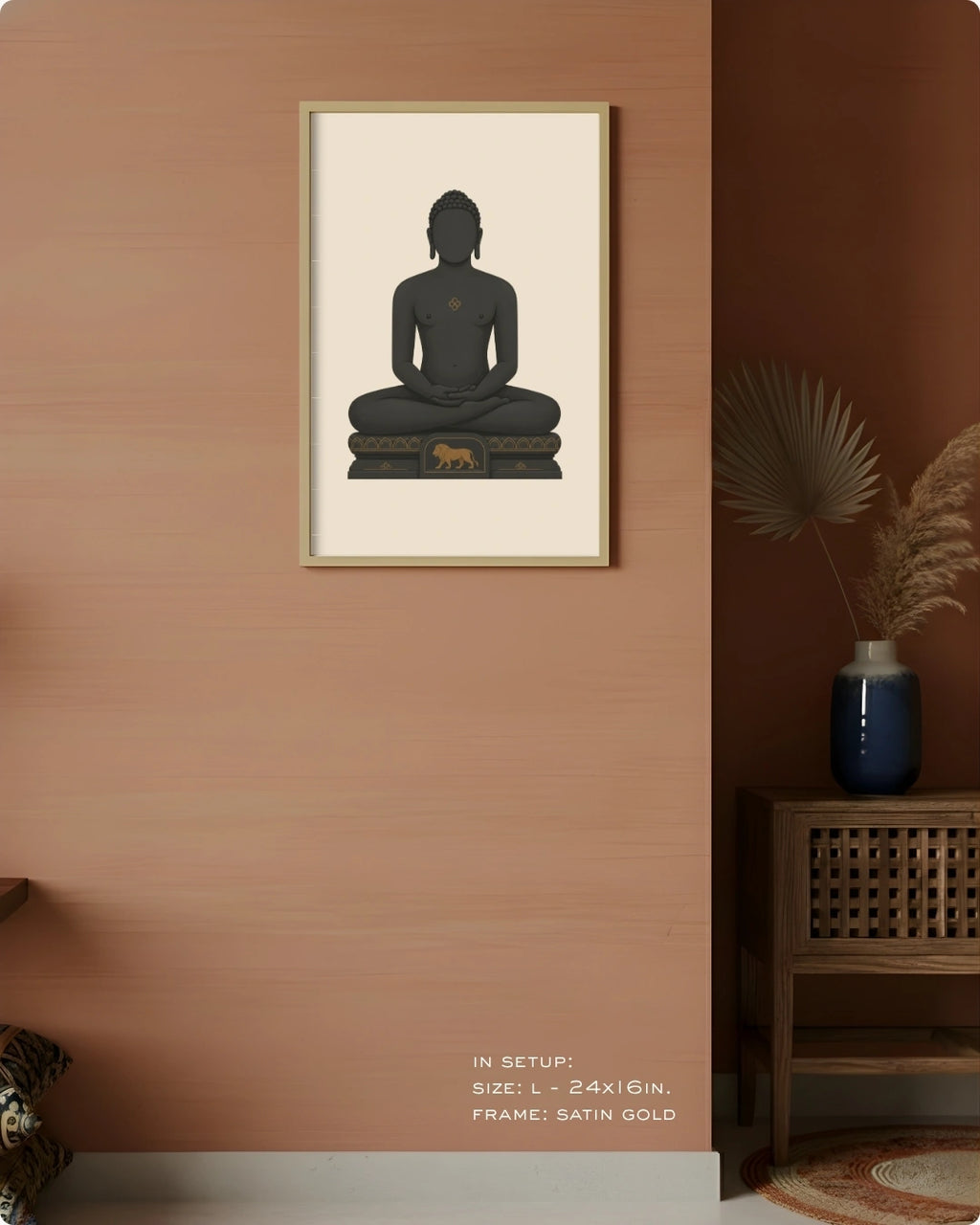A framed wall art print featuring a seated Mahavira silhouette in dark charcoal on a warm cream background, hung on a peach-toned wall in a styled interior with a wooden side table, blue vase, dried palm leaves, and a small animal figurine.