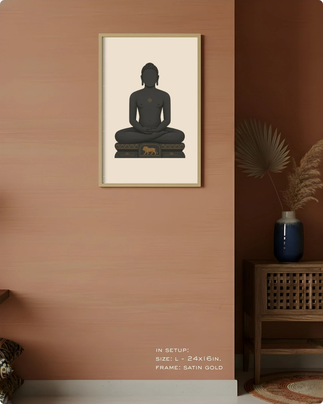 A framed wall art print featuring a seated Mahavira silhouette in dark charcoal on a warm cream background, hung on a peach-toned wall in a styled interior with a wooden side table, blue vase, dried palm leaves, and a small animal figurine.