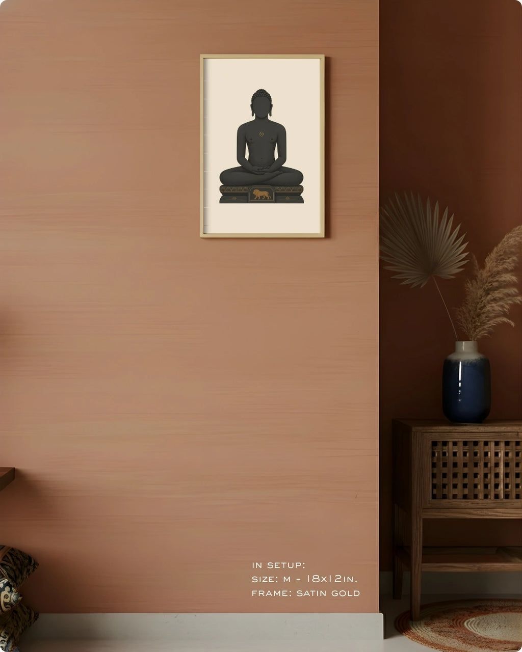 A framed wall art print showing a seated Mahavira silhouette in dark charcoal on a cream background, hung on a warm terracotta wall in a styled interior with a wooden side table, blue vase, dried palm leaves, and small text at the bottom indicating size and frame details.