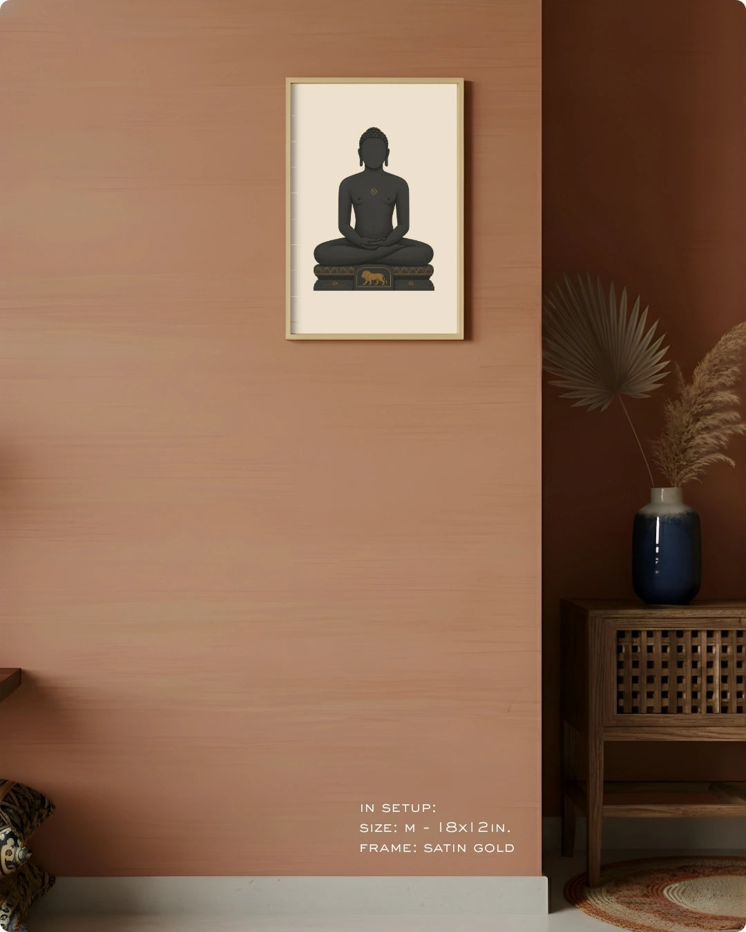 A framed wall art print showing a seated Mahavira silhouette in dark charcoal on a cream background, hung on a warm terracotta wall in a styled interior with a wooden side table, blue vase, dried palm leaves, and small text at the bottom indicating size and frame details.