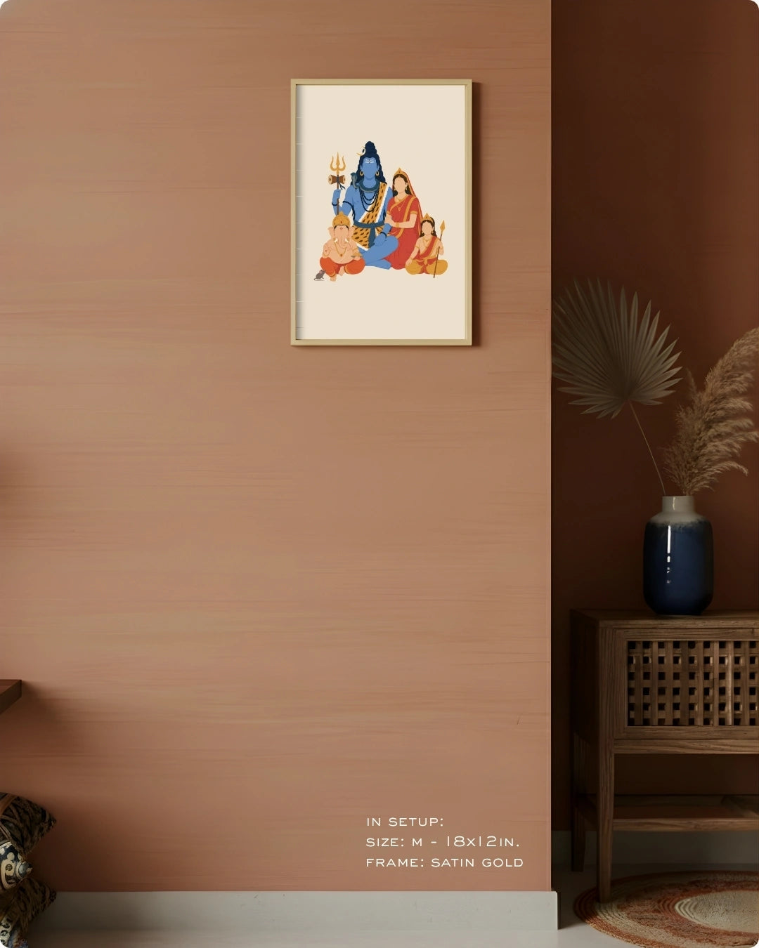 A framed art print depicting a traditional Indian religious scene with blue and red figures on a cream background, hung on a textured terracotta wall in a styled room setting with a wooden side table and vase.