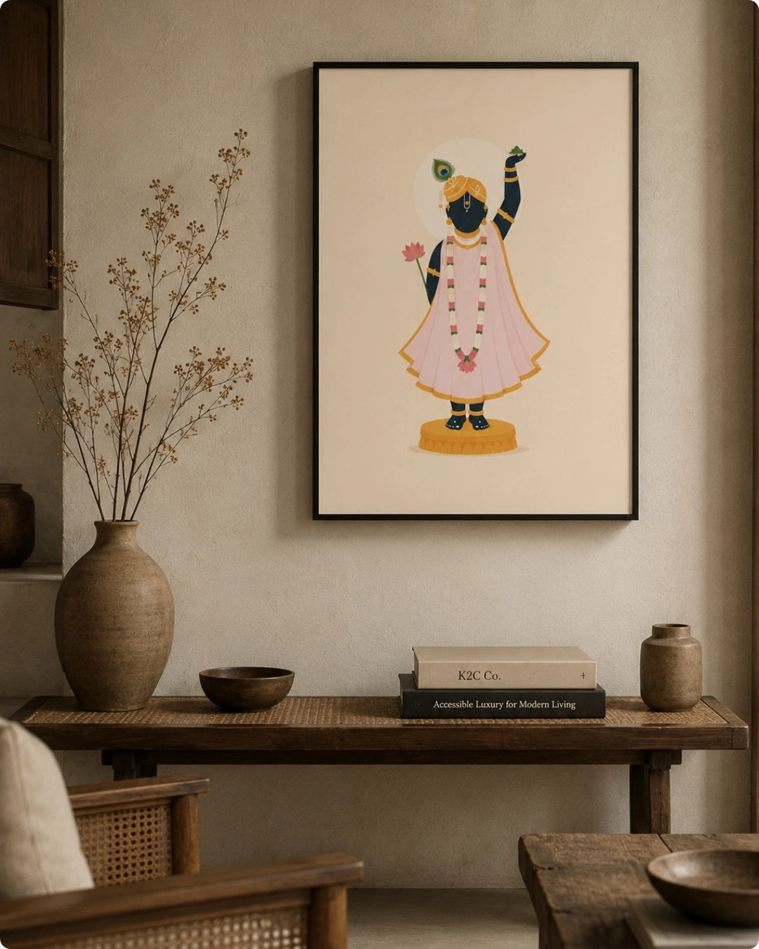 A framed art print featuring a stylized illustration of Shrinathji in a pink kurta and yellow turban, hanging on a textured beige wall above a wooden console table with decorative vases and books.