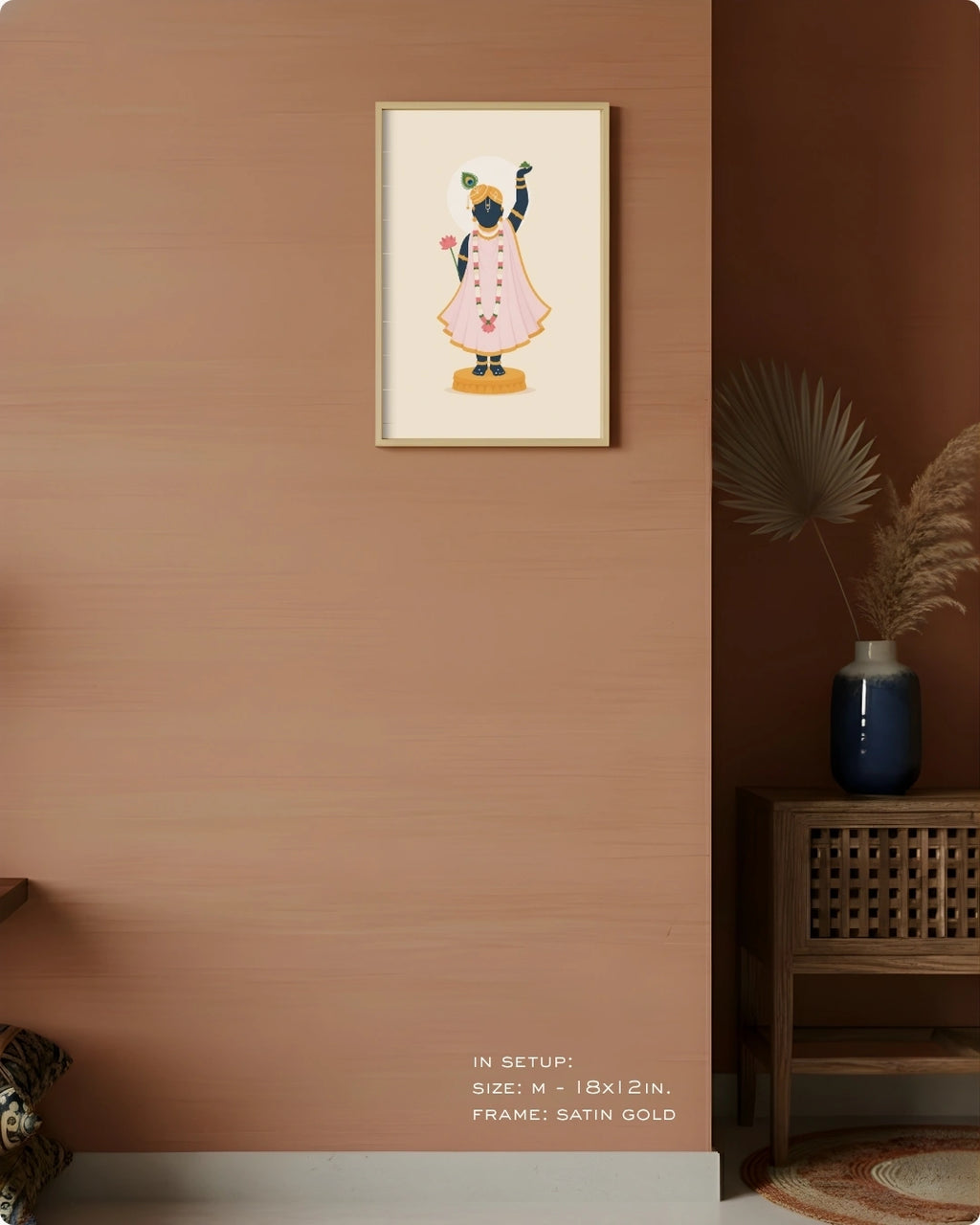 A framed art print featuring Shrinathji in a pink dress, displayed on a warm terracotta wall in a styled interior setting with a wooden side table and dried pampas grass.