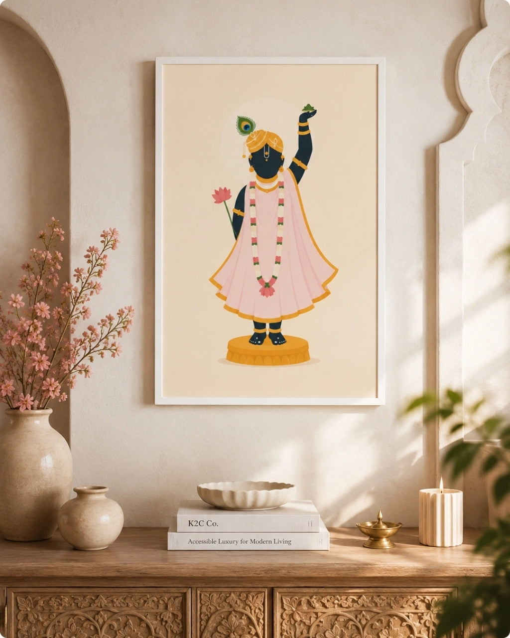 A framed art print featuring a stylized illustration of Lord Shrinathji in a pink kurta and yellow turban, hanging on a beige wall above a wooden console table decorated with vases, books, and a candle.