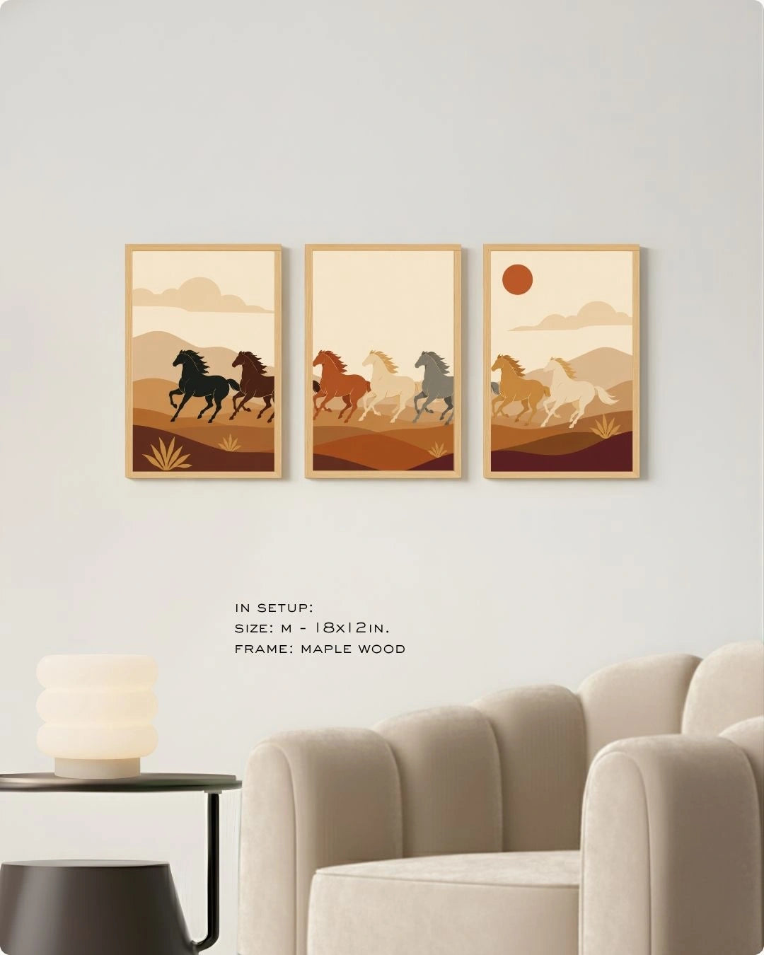 A set of three medium maple wood-framed wall art prints hanging above a plush cream velvet armchair in a modern minimalist lounge. The triptych features stylized horses running through an earth-toned desert landscape. Text on the wall indicates the size is M - 18x12 inches. A sleek black side table with a contemporary stacked frosted glass lamp sits next to the chair.