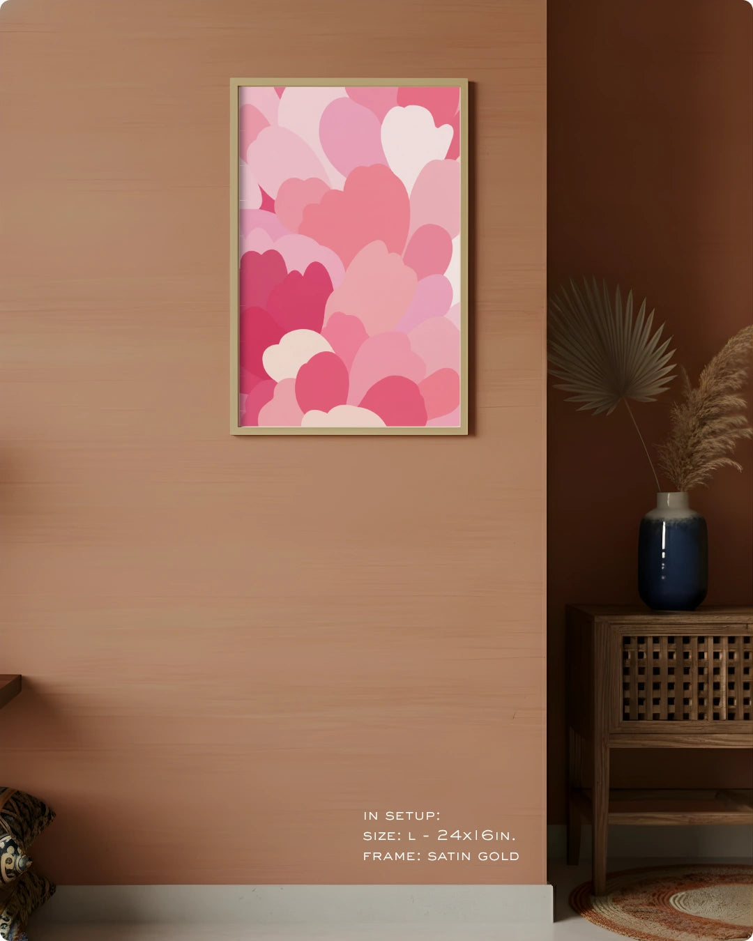 A framed wall art print featuring overlapping hearts in shades of pink, coral, and cream, displayed on a warm peach-colored wall in a styled interior setting with a wooden side table and vase.