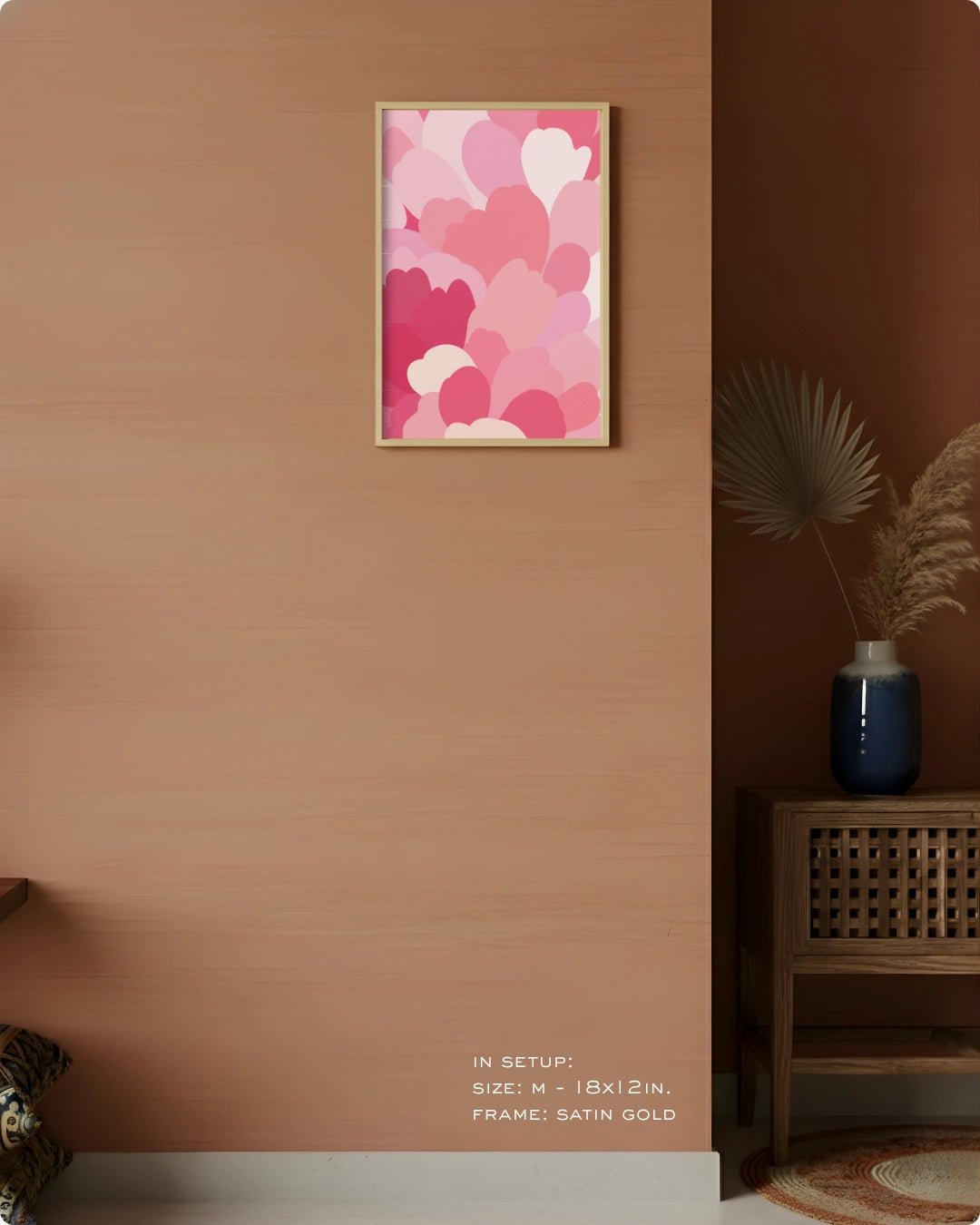 A framed wall art print featuring overlapping pink, red, and white hearts, displayed on a warm terracotta wall in a styled interior setting with a wooden side table, blue vase, and dried palm leaves.