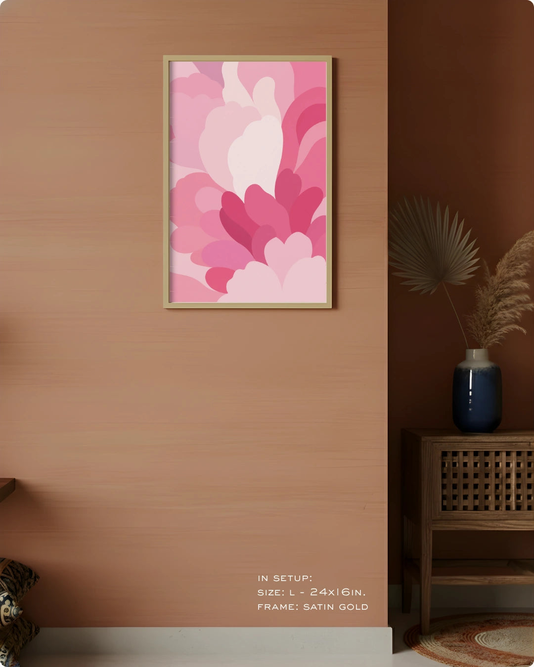 A framed abstract floral wall art print in pink tones hangs on a warm peach wall in a styled interior setting. The artwork features layered petal-like shapes in shades of pink, magenta, and light blush.