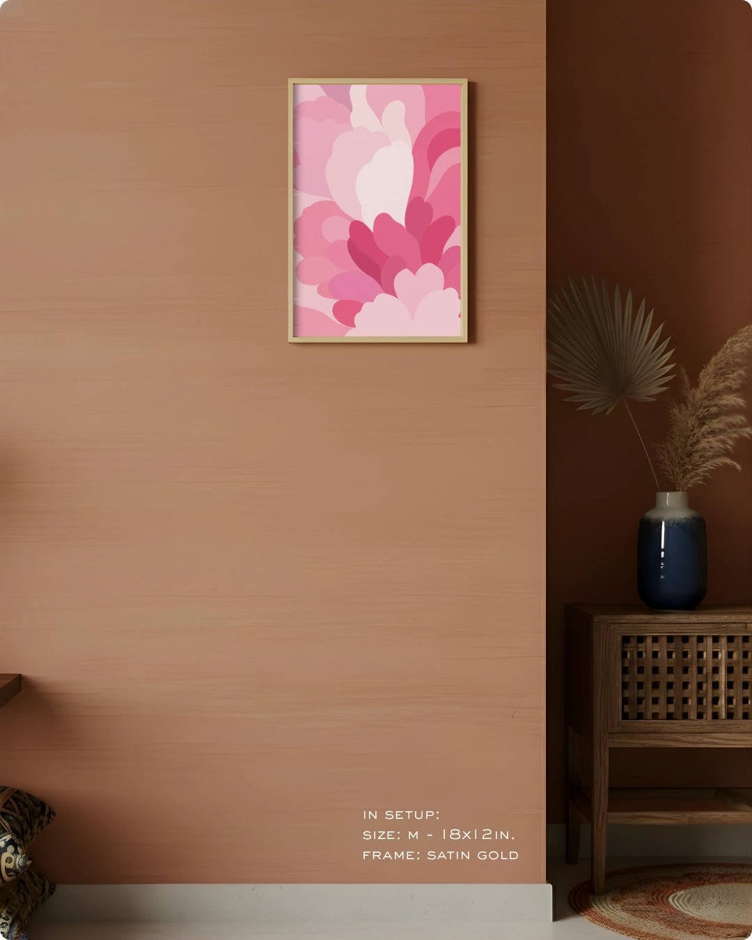 A framed wall art print featuring layered pink and white abstract floral shapes, displayed on a warm terracotta wall in a styled interior setting with a wooden side table, blue vase, dried palm leaves, and small text at the bottom indicating size and frame details.