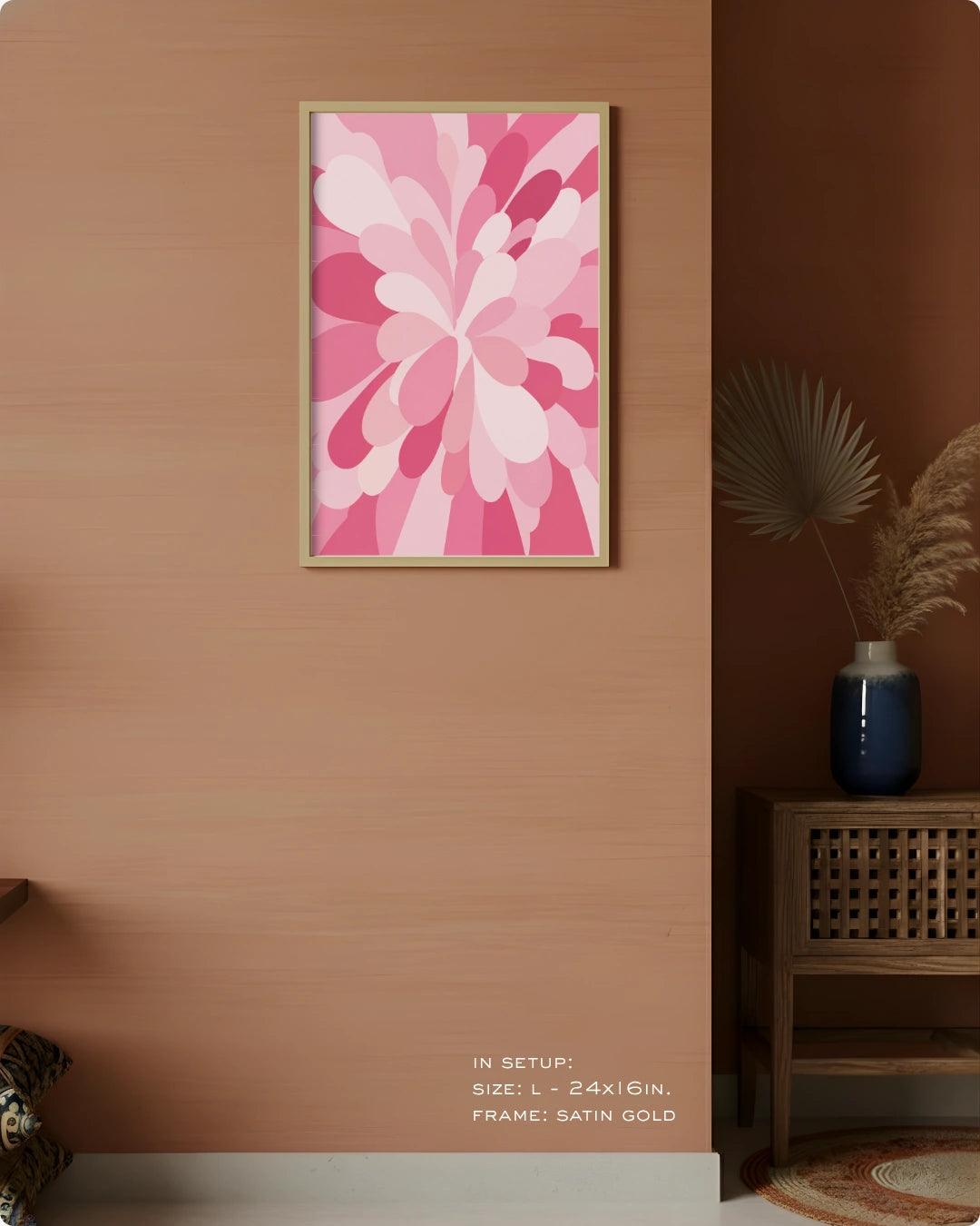 A framed abstract floral wall art print in pink and white tones hangs on a warm peach wall in a styled interior setting with a wooden side table, blue vase, dried palm leaves, and a small rug. Text at the bottom reads: 'IN SETUP: SIZE L = 24x16in.