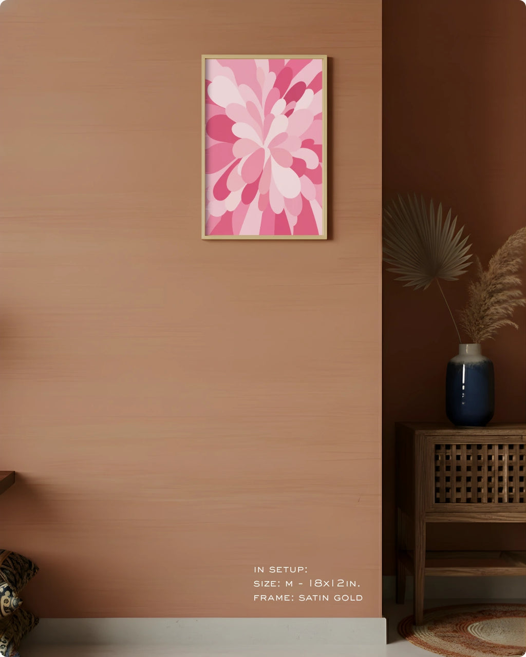 A framed wall art print featuring a pink and white floral design hanging on a warm terracotta-colored wall in a styled interior setting with a wooden side table, blue vase, and dried palm leaves.