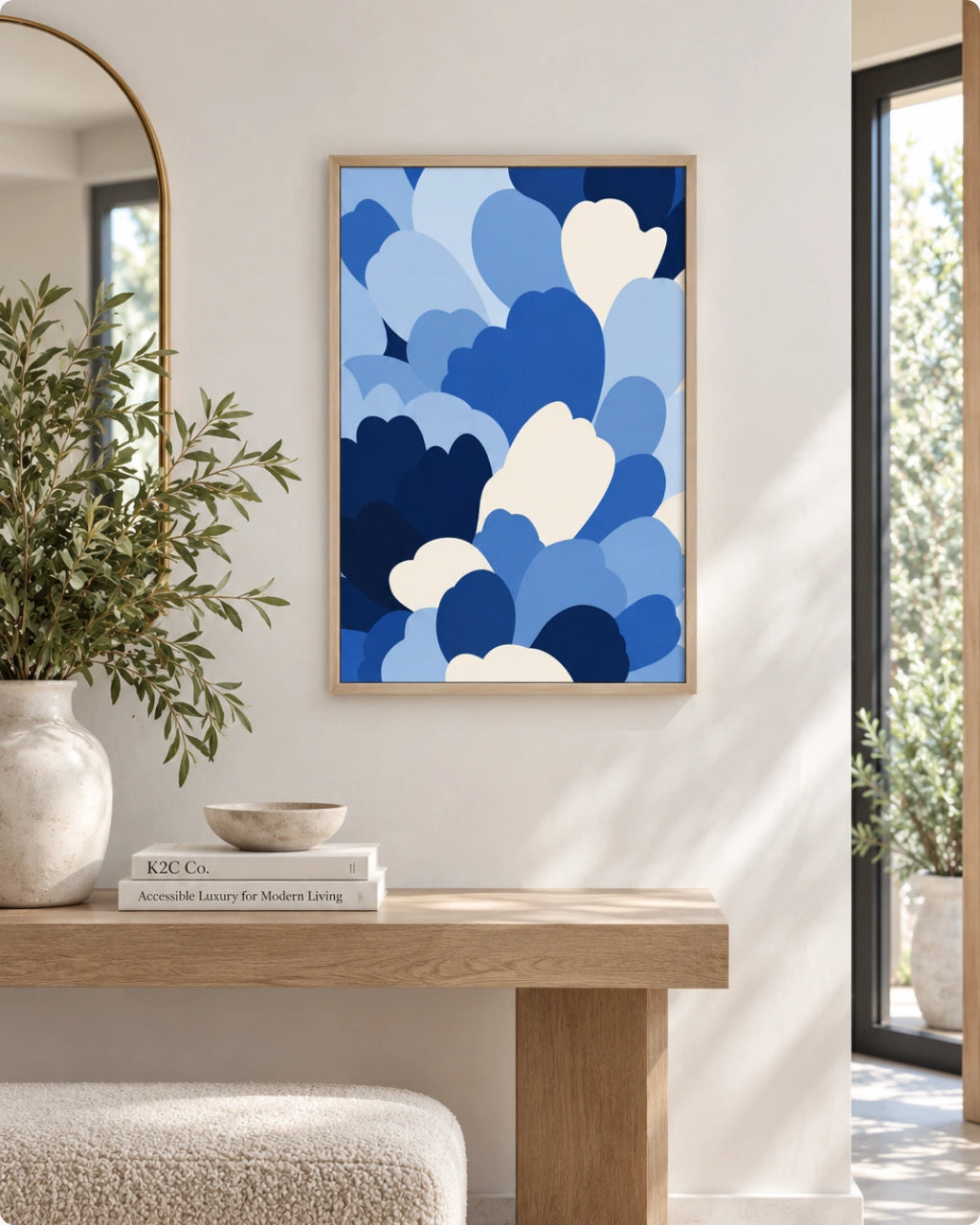 A framed abstract art print featuring overlapping blue and white cloud-like shapes, displayed on a white wall above a light wood console table in a bright, modern interior.