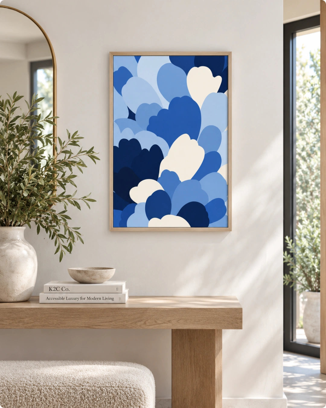 A framed abstract art print featuring overlapping blue and white cloud-like shapes, displayed on a white wall above a light wood console table in a bright, modern interior.