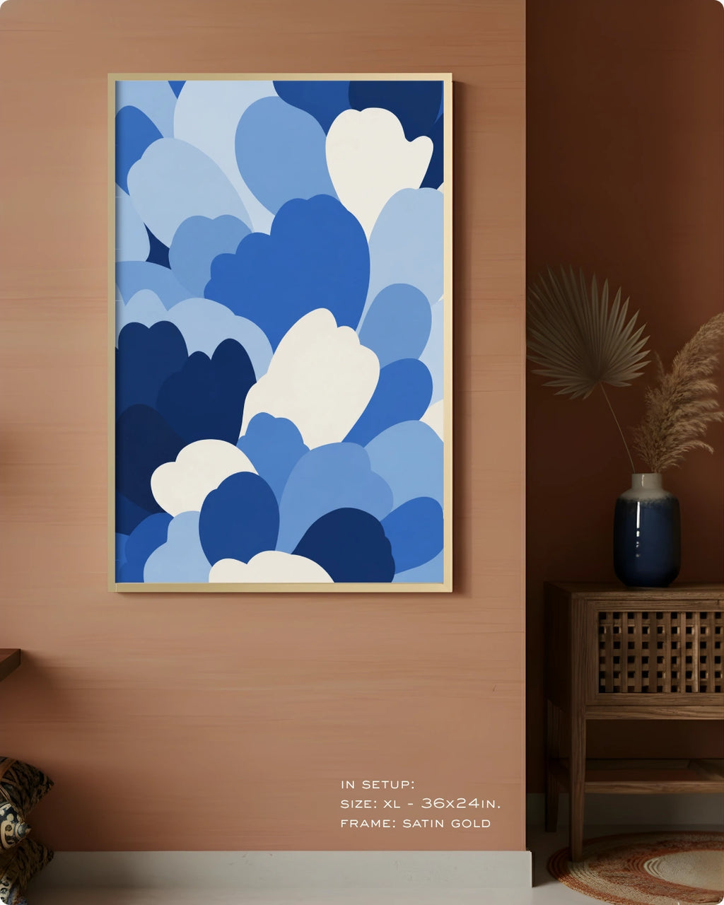 A framed abstract wall art print with layered blue and white cloud-like shapes, displayed on a warm peach wall in a styled interior setting with a side table and vase.