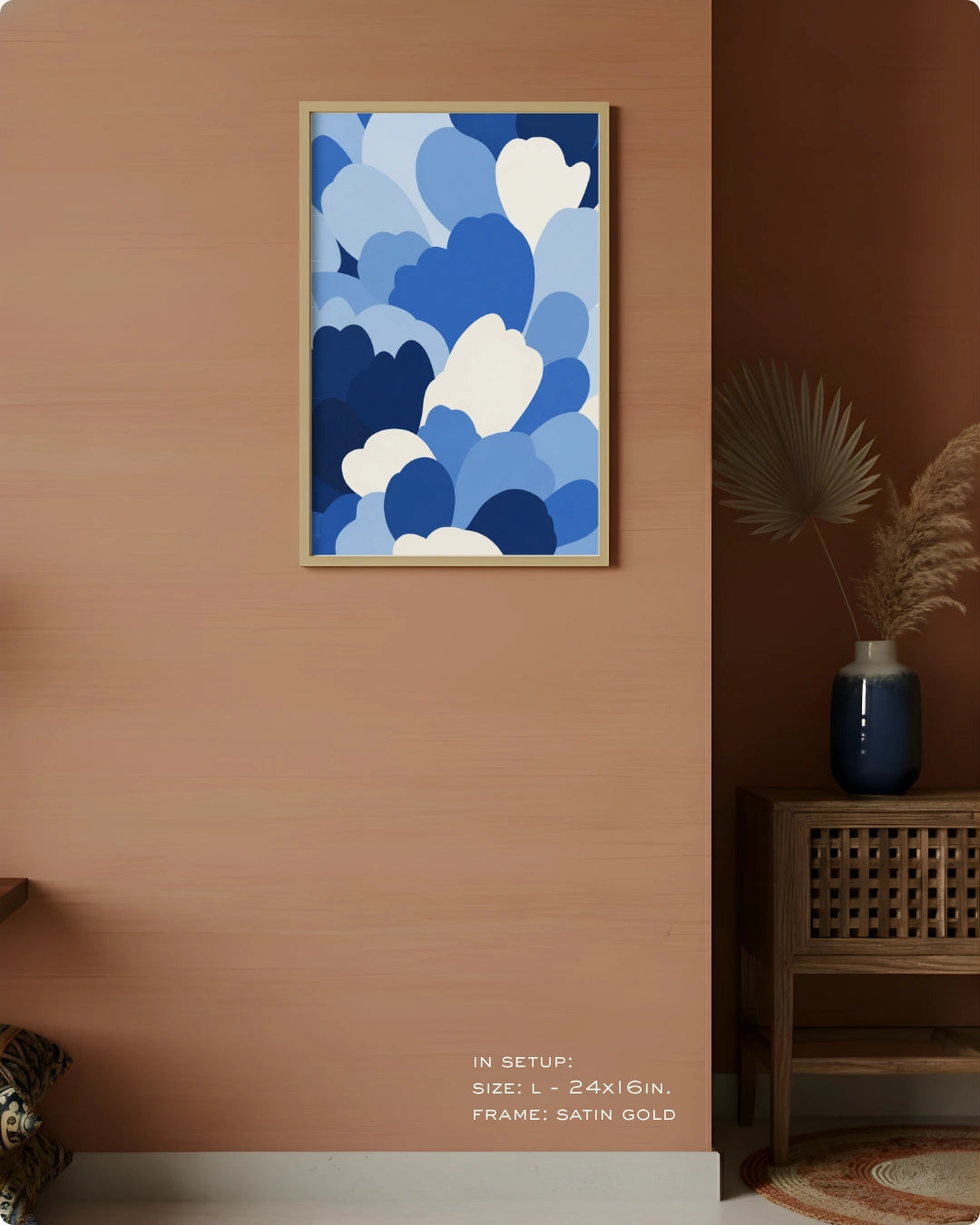 A framed abstract art print featuring blue and white cloud-like shapes, displayed on a warm terracotta wall in a styled interior setting with a side table and vase.