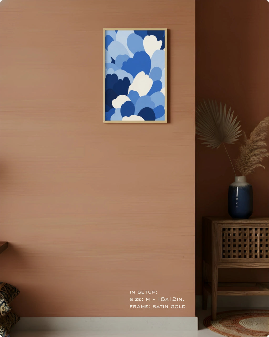 A framed abstract art print featuring blue and white cloud-like shapes hanging on a warm terracotta wall in a styled interior setting with a wooden side table, blue vase, and dried palm leaves.