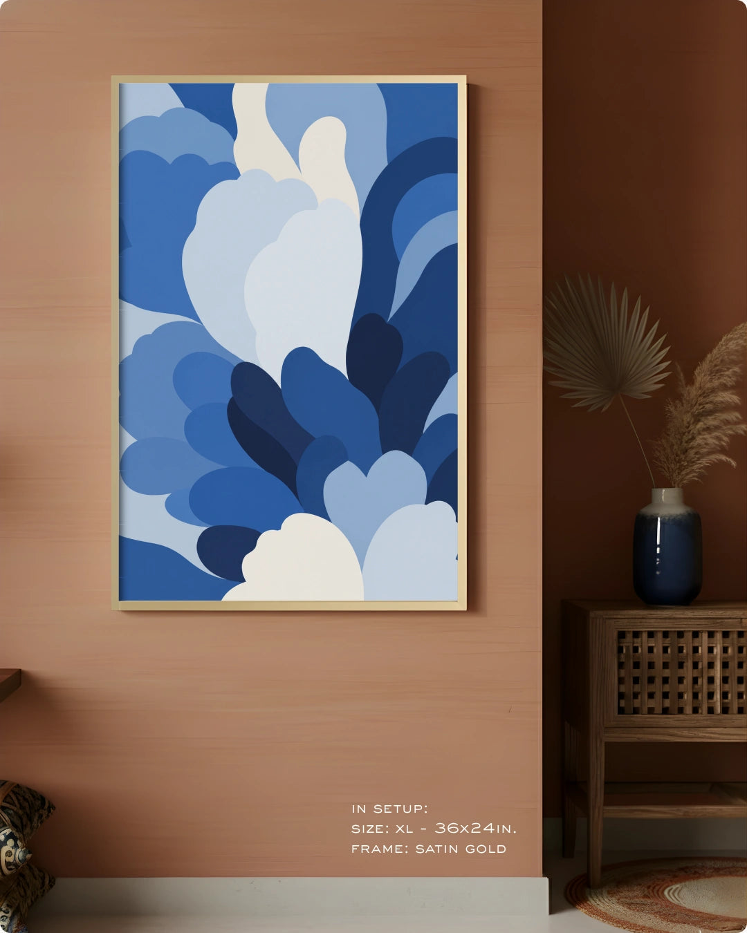 A framed abstract wall art print with layered blue and white organic shapes hanging on a warm terracotta wall in a styled interior. The frame appears satin gold.